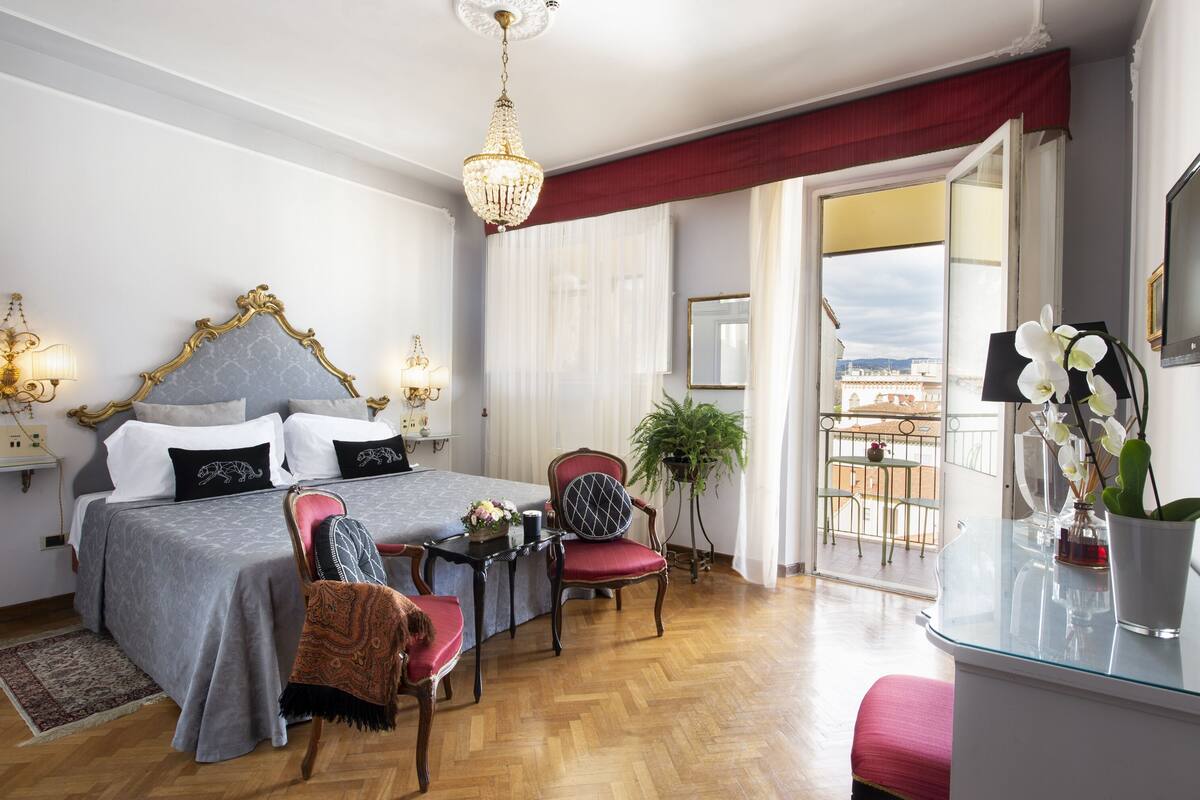 A hotel room with a large bed, a sitting area with a table and chairs, a balcony with a view, and a chandelier.