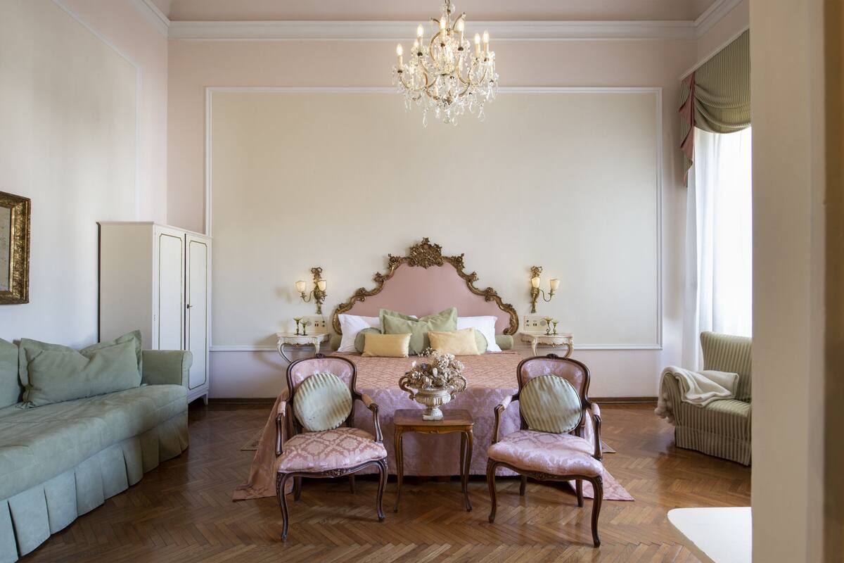 A bedroom with a large bed, two chairs, a small table, and a chandelier.