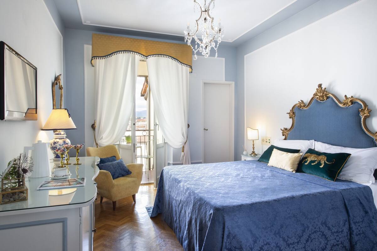 A bedroom with a large bed, a chair, a desk with a lamp, and a chandelier.