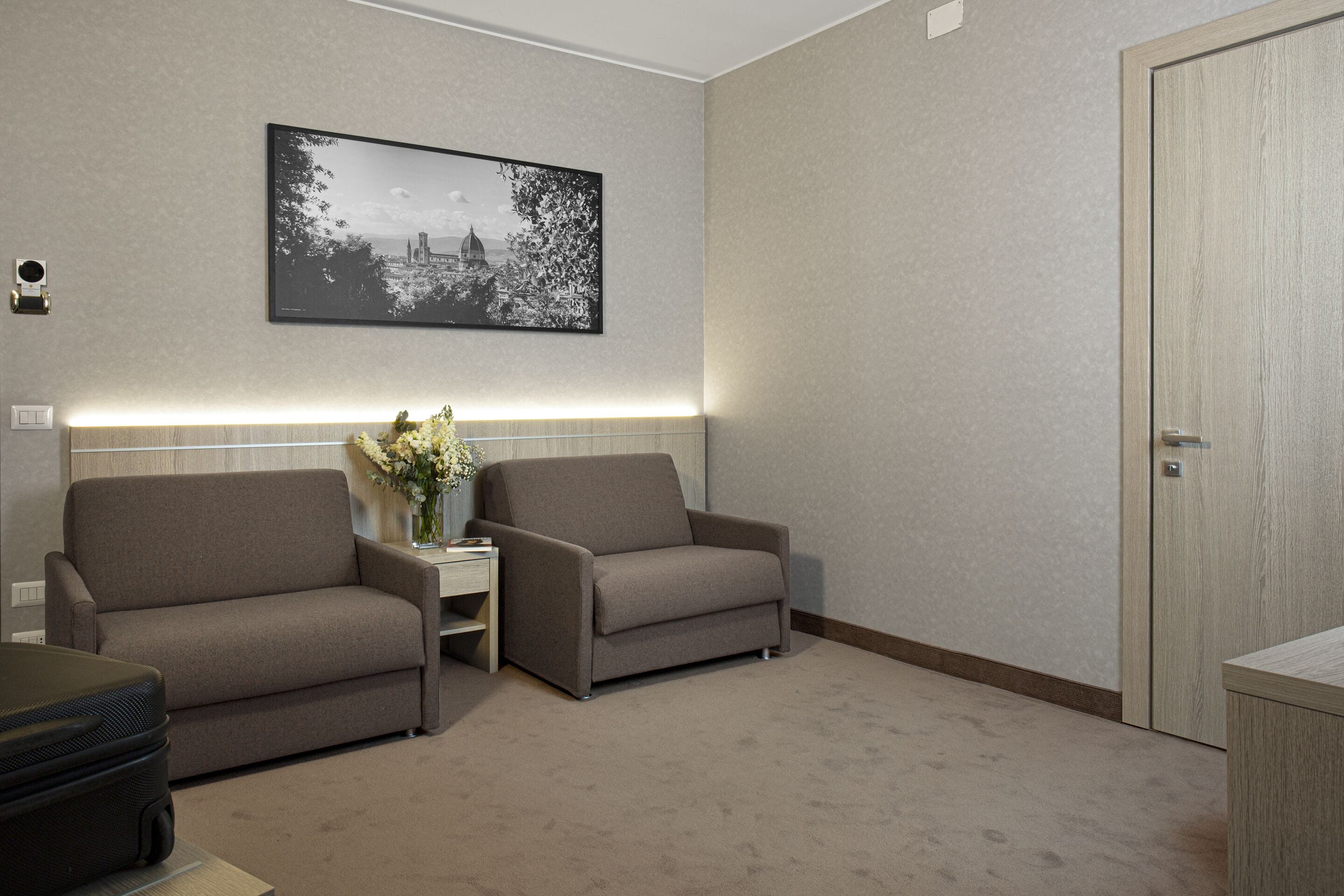 A hotel room with two sofas, a small table with a vase of flowers, a framed picture on the wall, and a door.