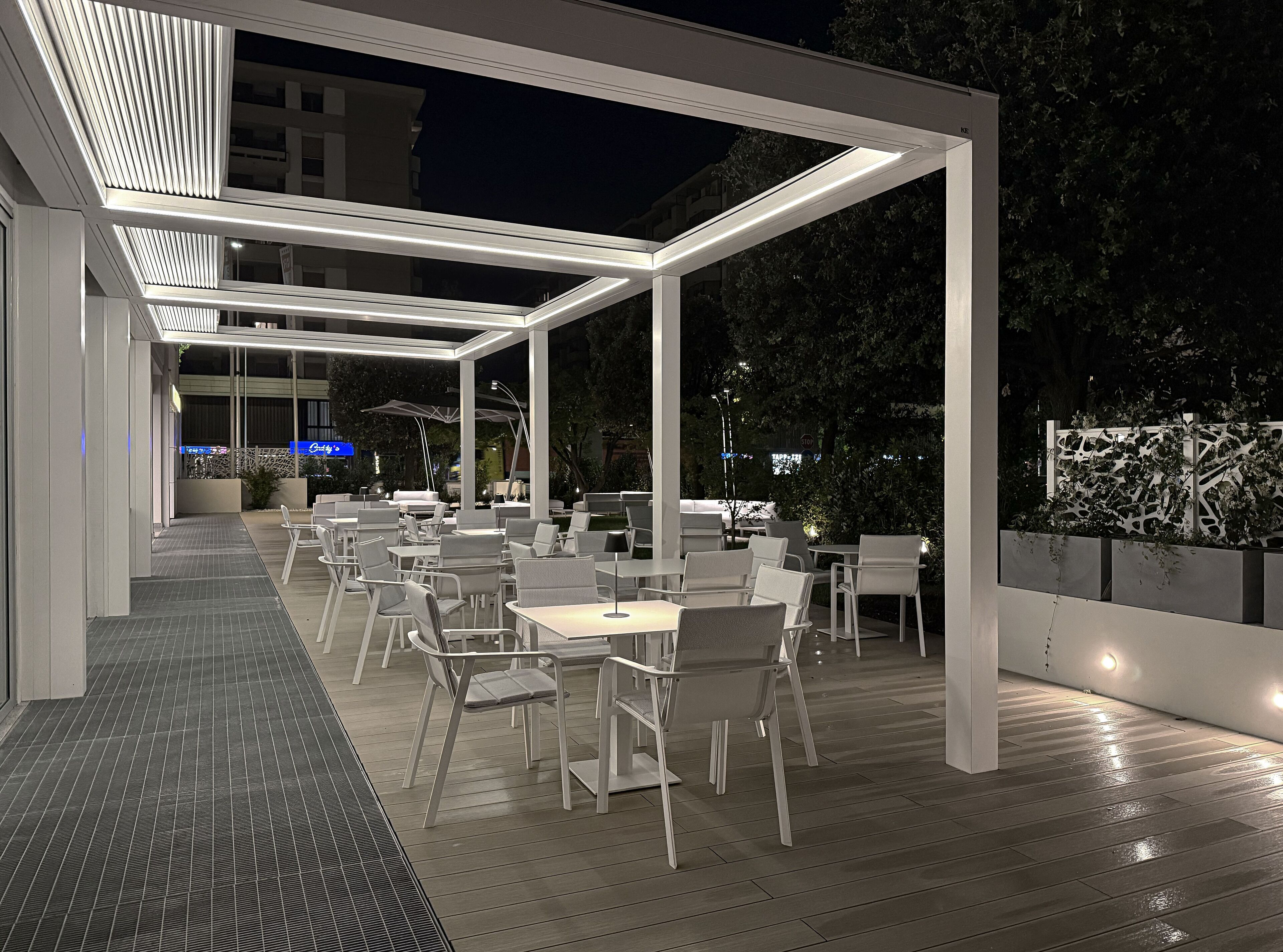 An outdoor patio area with white chairs and tables, a covered walkway, and a view of the surrounding area at night.