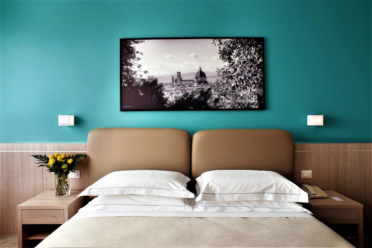A hotel room with a bed, a nightstand with a vase of flowers, and a wall-mounted picture of a cityscape.