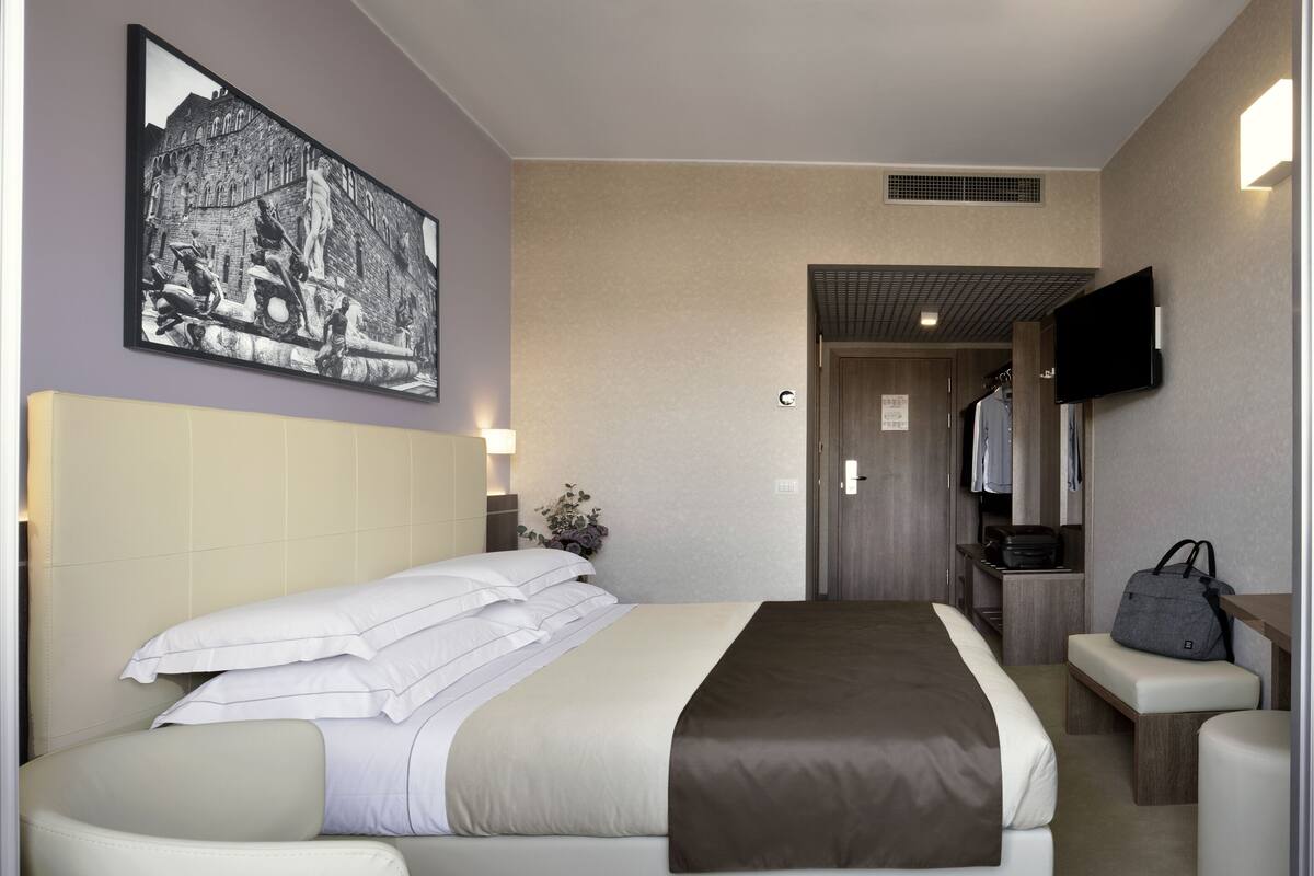 A hotel room with a bed, a chair, a desk, a TV, and a painting on the wall.