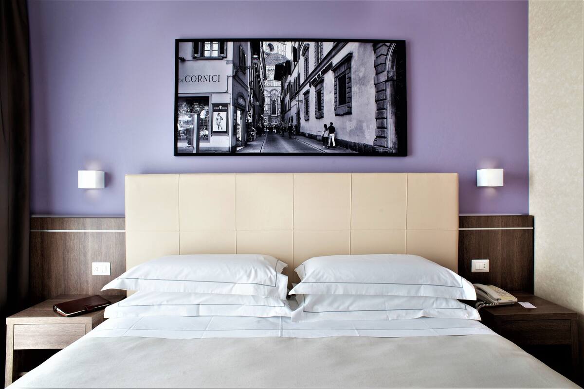 A hotel room with a large bed, a headboard, bedside tables, a nightstand, a wall-mounted picture, and a wall socket.