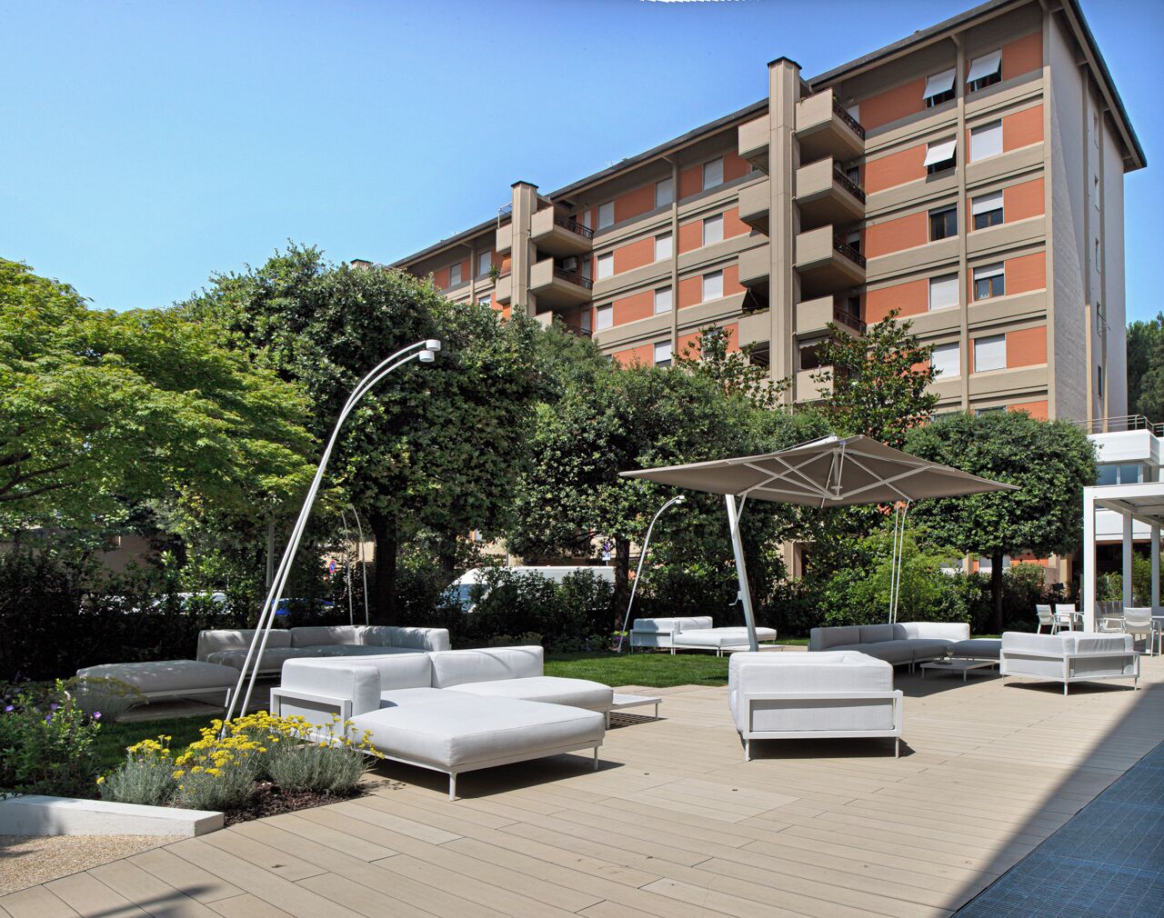 Un área de lounge al aire libre, moderna, con sofás blancos y sombrillas, rodeada de follaje y un edificio de varios pisos al fondo.