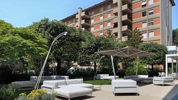 Un área de lounge al aire libre, moderna, con sofás blancos y sombrillas, rodeada de follaje y un edificio de varios pisos al fondo.