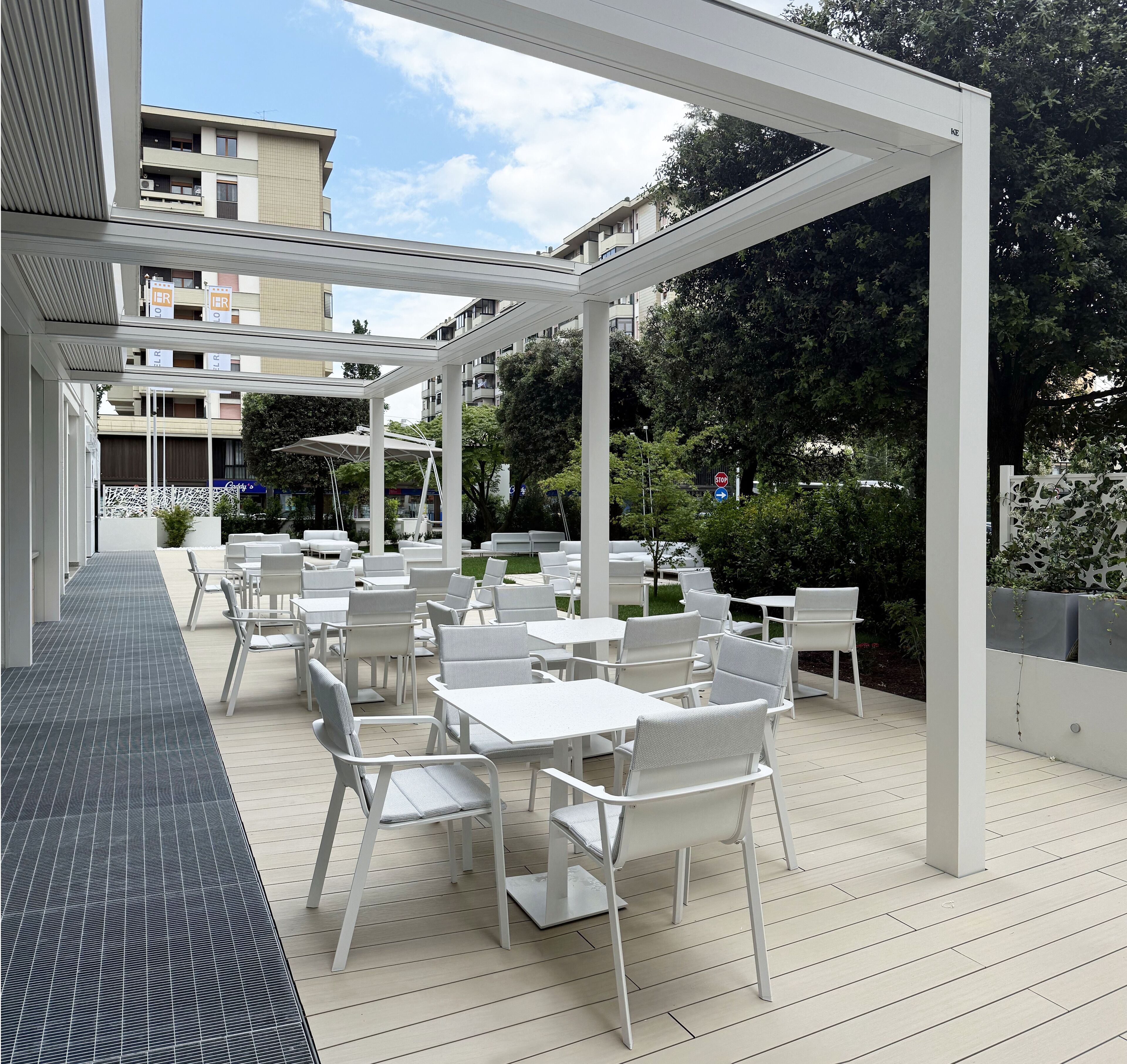 Un patio al aire libre con mesas y sillas blancas, un área de estar techada y vistas a edificios y vegetación circundante.
