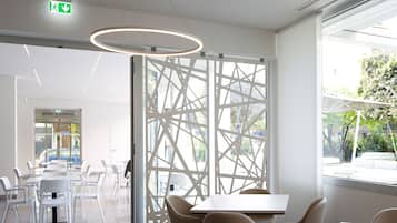 Ein modernes Interieur mit Essbereich, ausgestattet mit weißen Tischen und Stühlen, einer geometrischen Glasscheibe und Blick ins Freie.