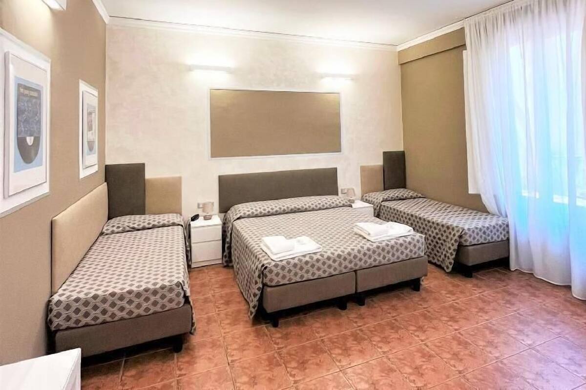 Une chambre d’hôtel avec trois lits, une grande fenêtre avec des rideaux et un luminaire fixé au mur.
