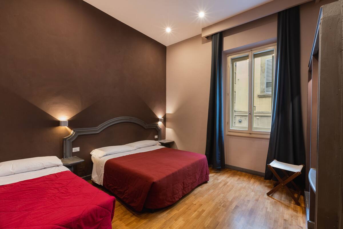 A hotel room with two beds, wooden flooring, a window with curtains, and wall-mounted lights.