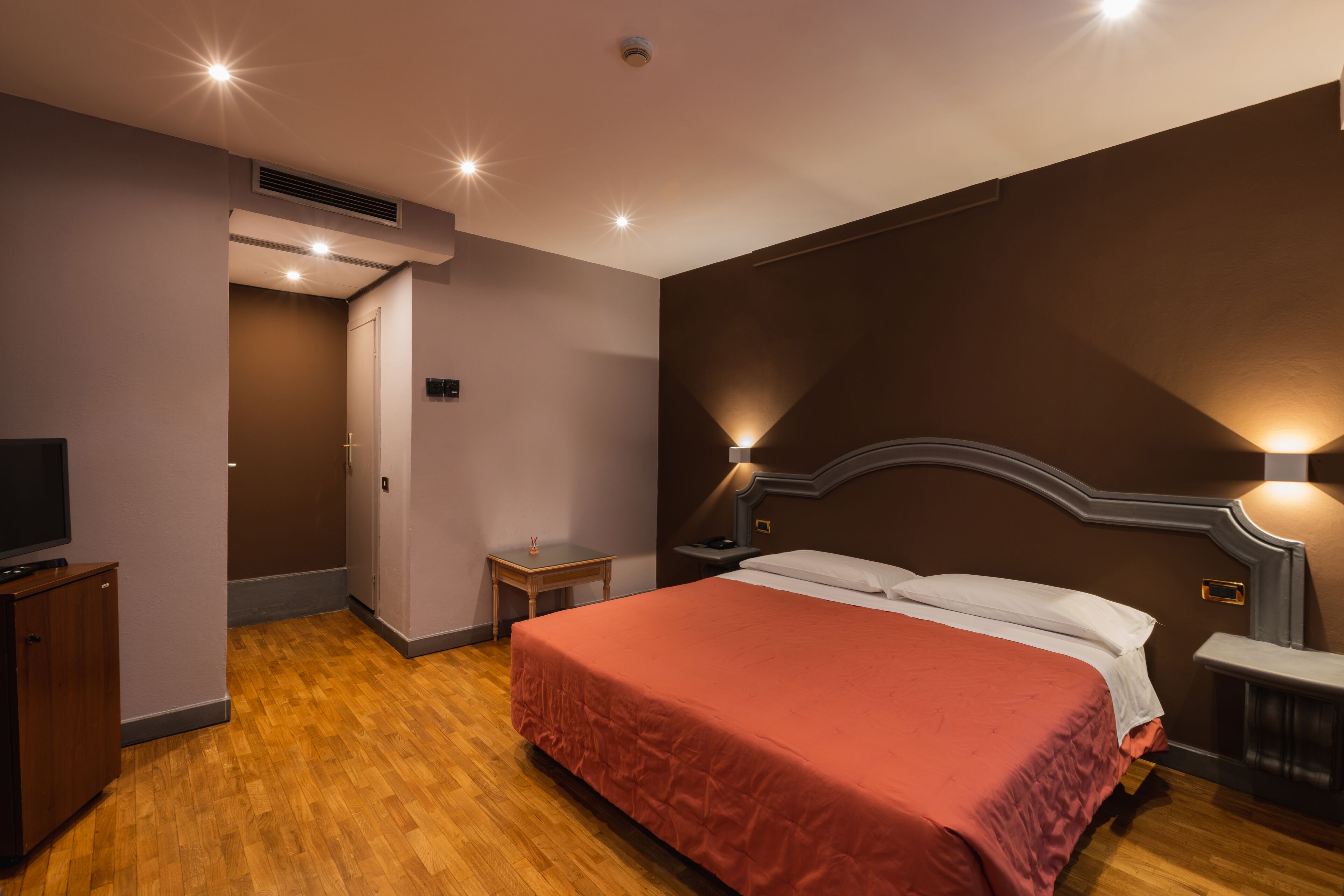 A bedroom with a wooden floor, a bed with a red bedspread, a nightstand, a TV on a cabinet, and a wall-mounted light fixture.