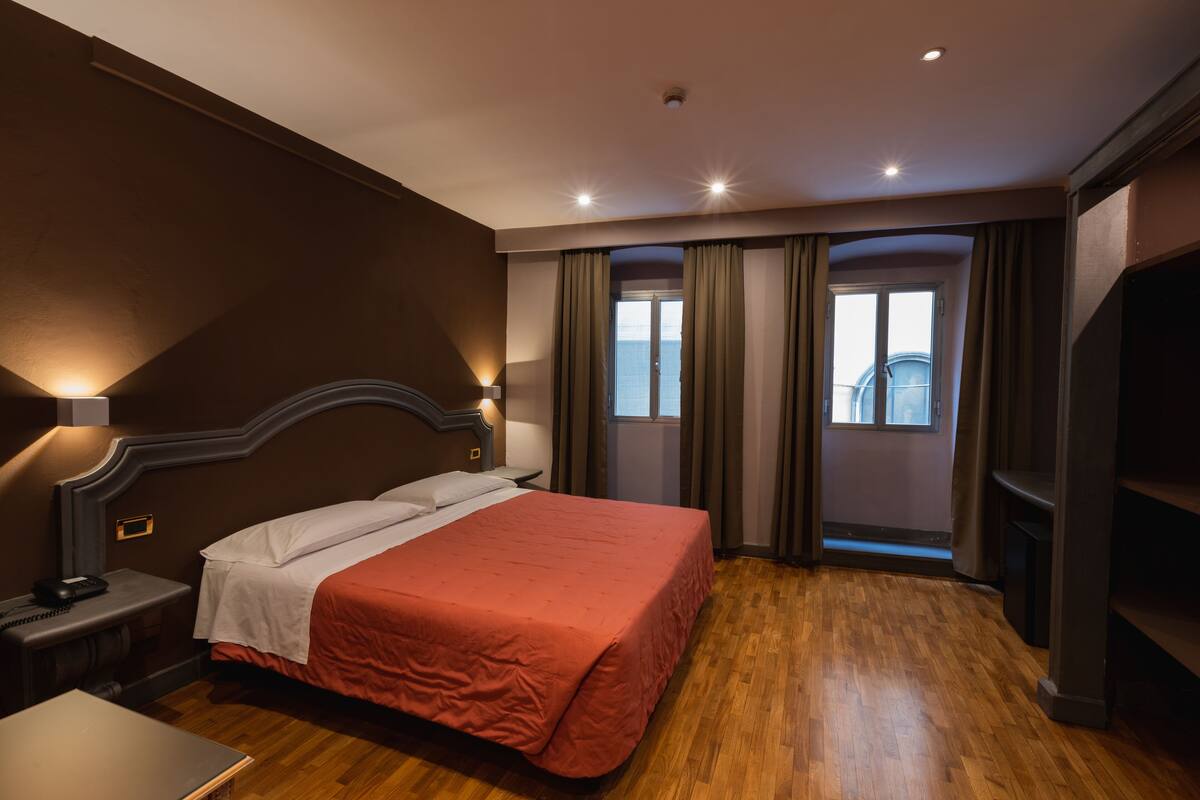 A hotel room with a large bed, a bedside table with a lamp, a wooden floor, and a view of the city through the window.