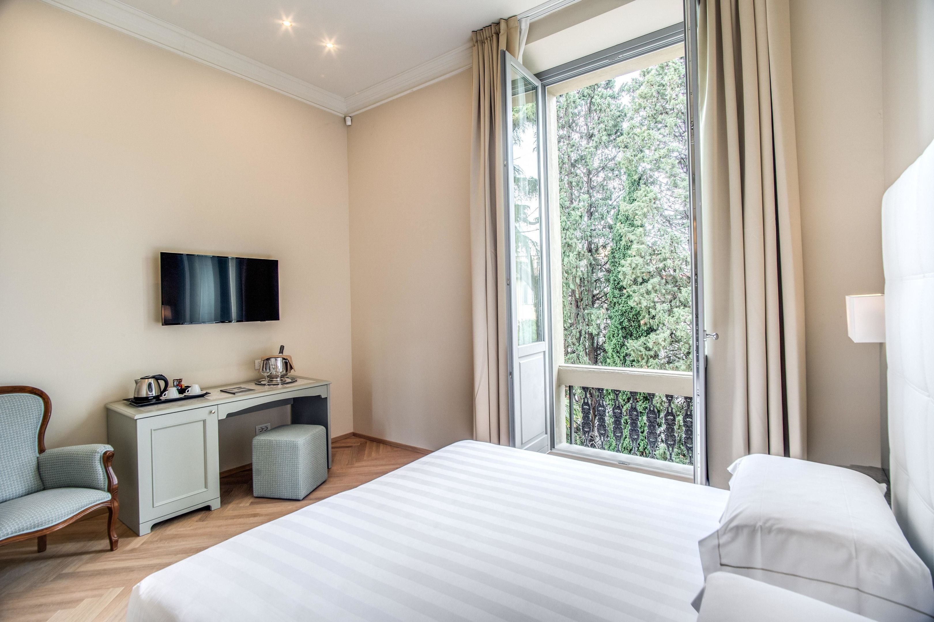 A hotel room with a bed, a desk with a flat-screen TV, a chair, and a window with curtains.