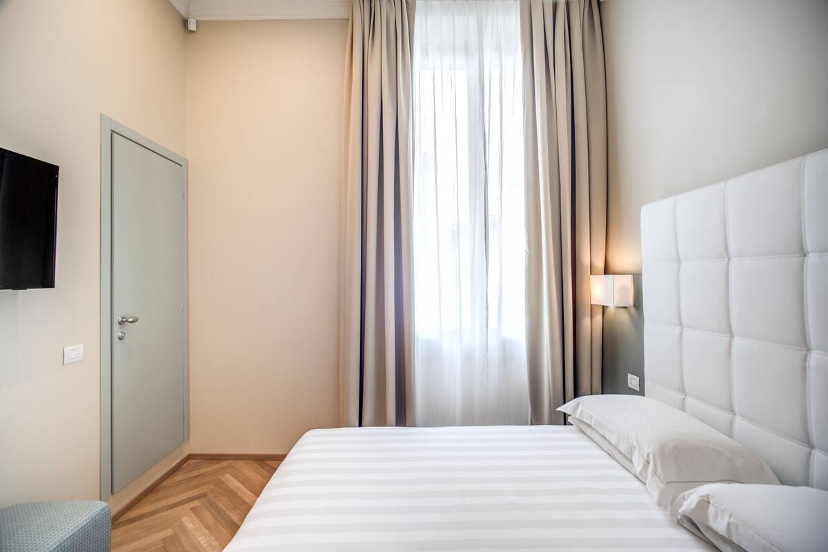 Habitación de hotel con cama, televisor montado en la pared, armario y ventana con cortinas.