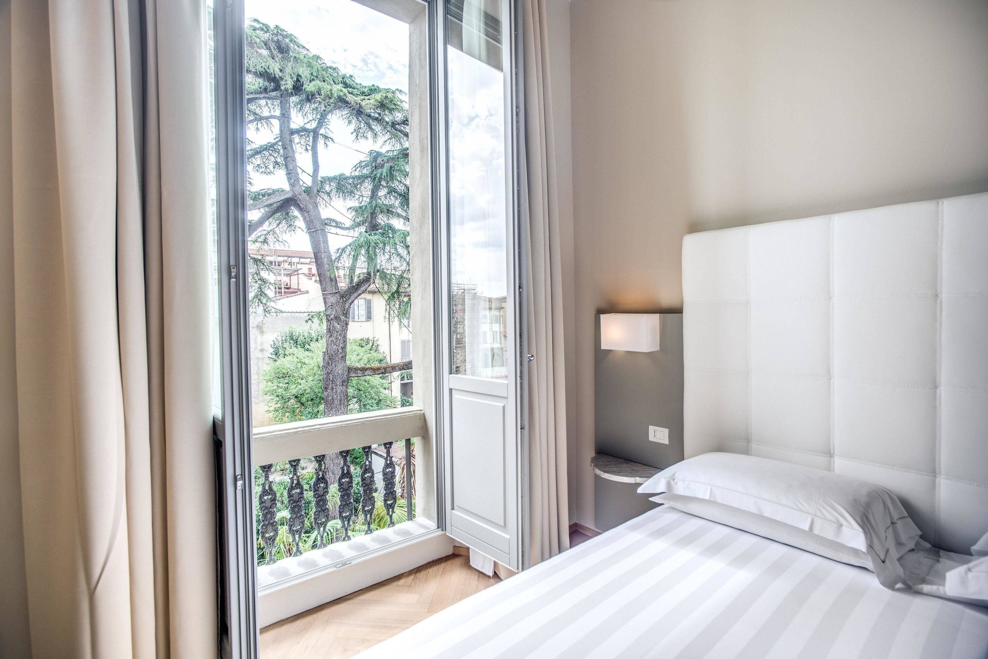 Une chambre avec une grande fenêtre, un lit recouvert de draps blancs, une table de chevet avec une lampe, et une vue sur la verdure à l’extérieur.
