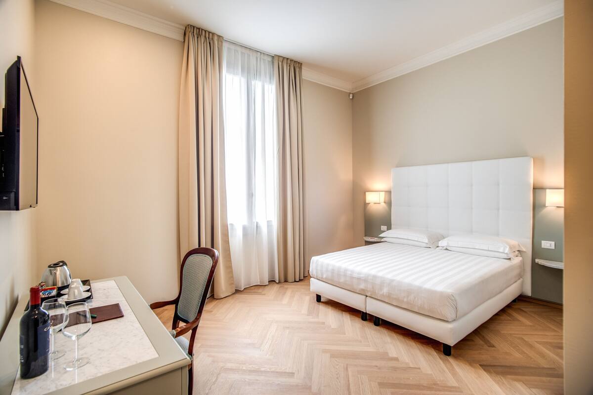 Habitación de hotel con cama, mesitas de noche, escritorio con silla, televisor y ventana con cortinas.