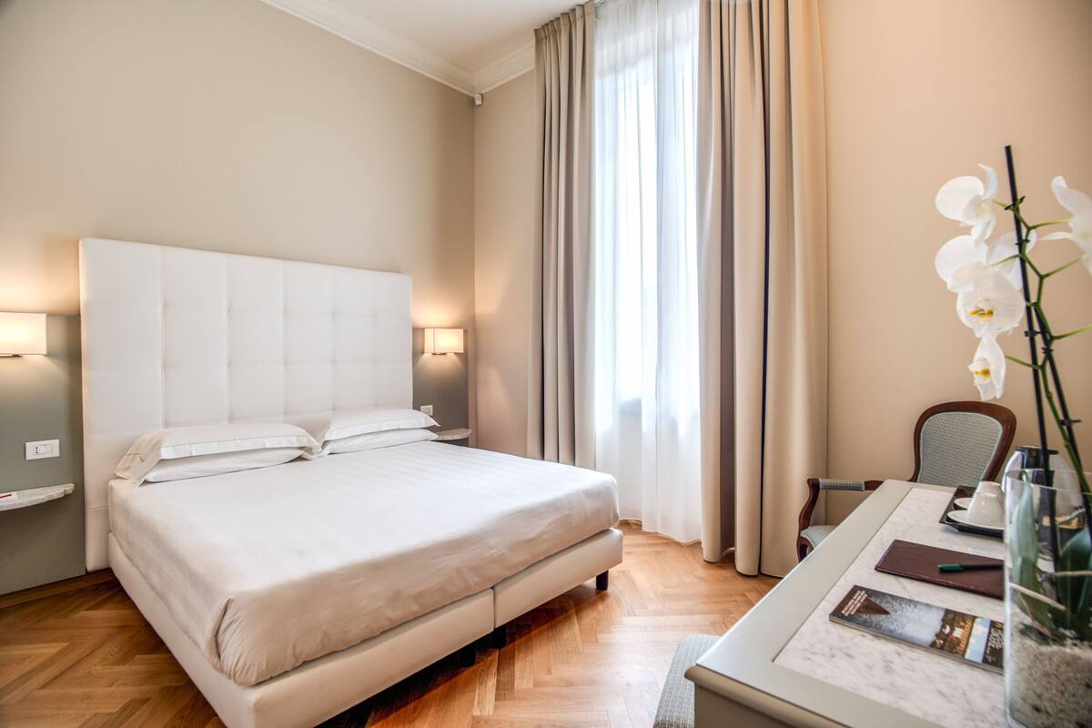 Una habitación de hotel con una cama grande, un escritorio con silla, un jarrón con flores y una ventana con cortinas.
