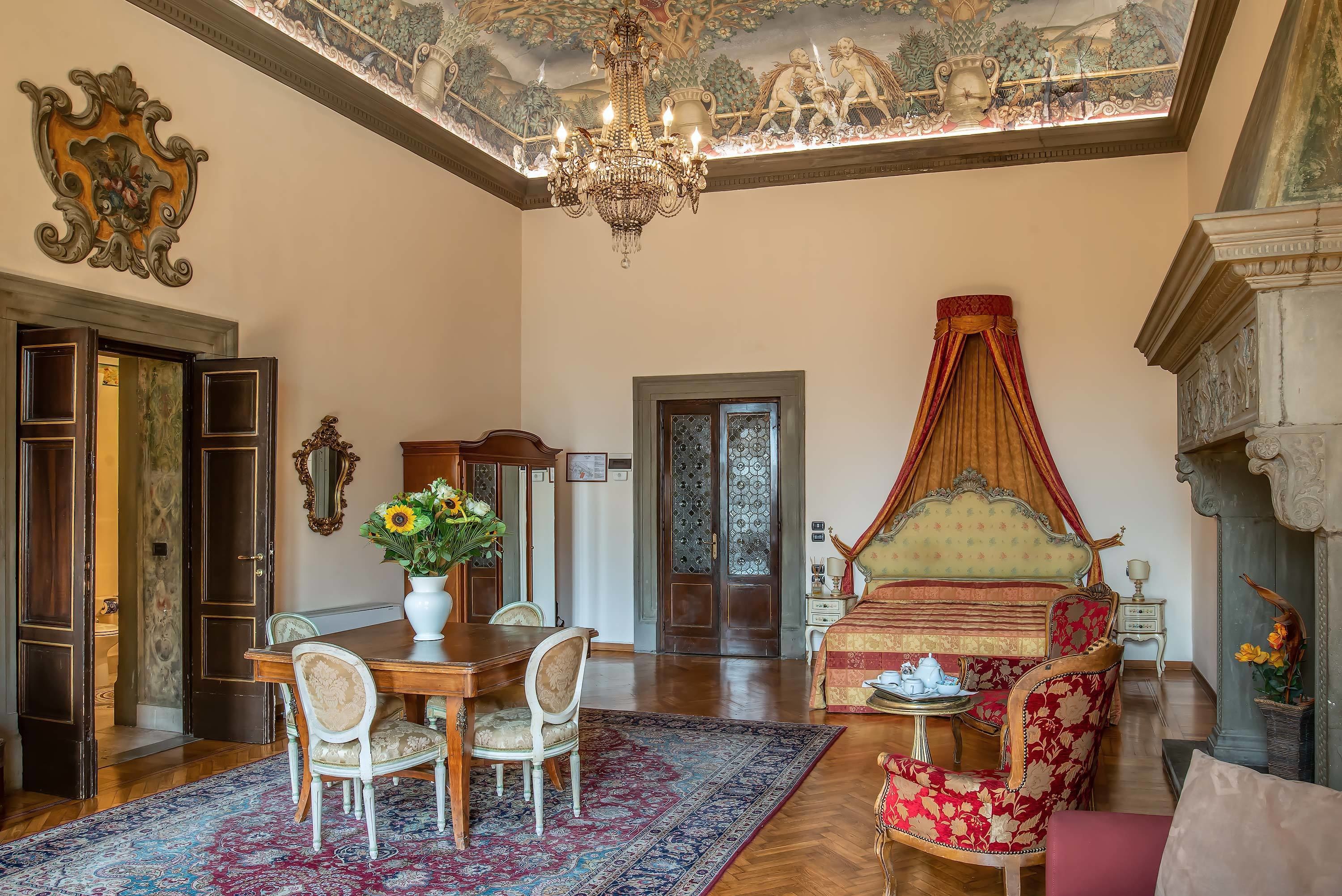 A spacious room with a large chandelier, a four-poster bed, a dining area with a table and chairs, and a detailed ceiling mural.