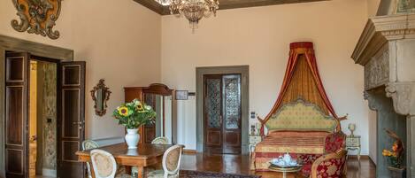 A spacious room with a large chandelier, a four-poster bed, a dining area with a table and chairs, and a detailed ceiling mural.
