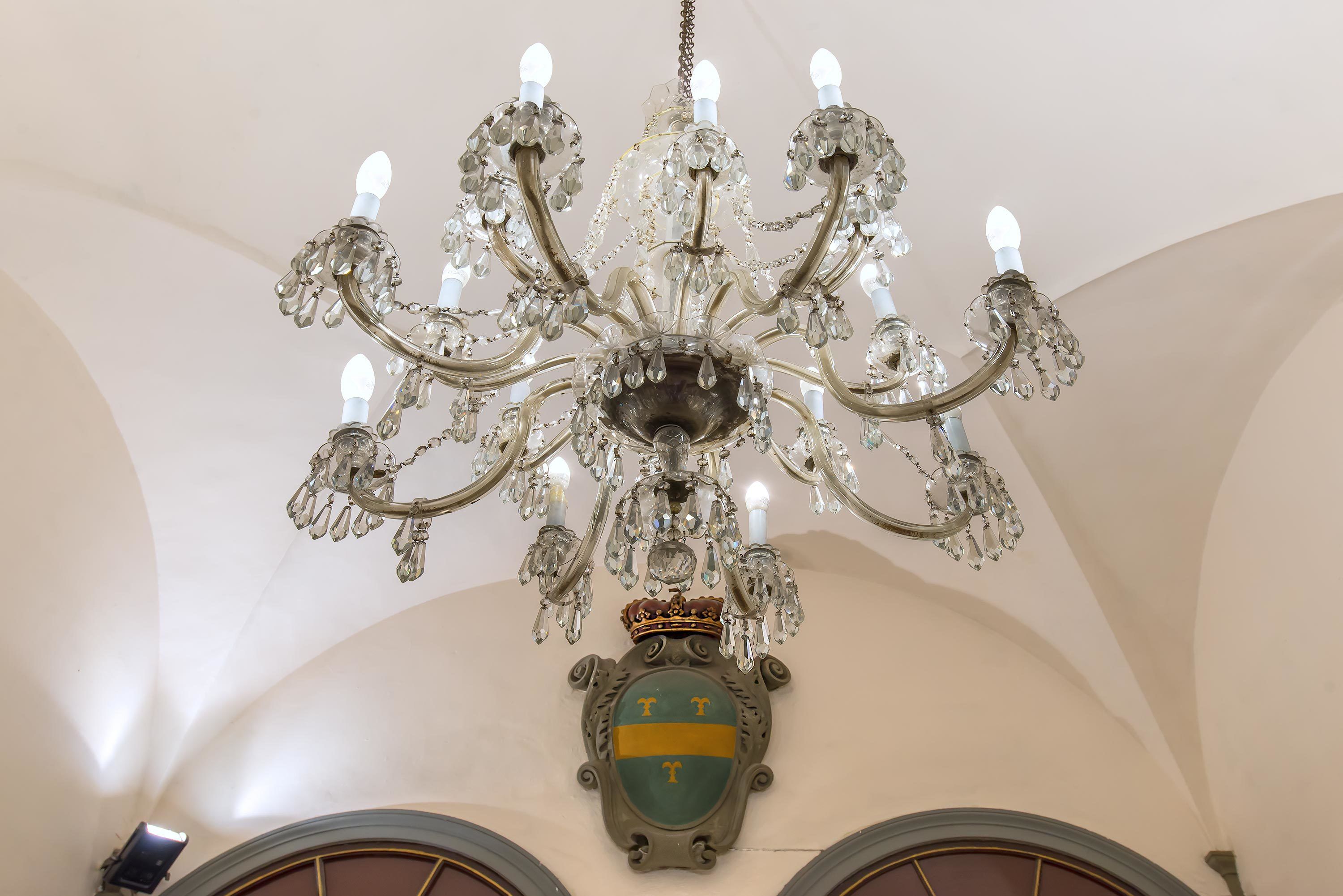 Ein Kronleuchter mit mehreren Leuchtarme und Kristallverzierungen, darunter ein Wappen, und gewölbte Decken.