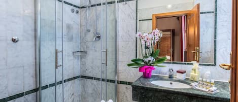 A modern bathroom with a glass-enclosed shower, a marble-tiled wall, a sink with toiletries, and a mirror reflecting the room.