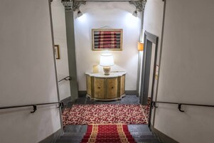 Una scala con una tappezzeria rossa a motivi decorativi, un armadio in legno con una lampada e un arazzo incorniciato al muro.
