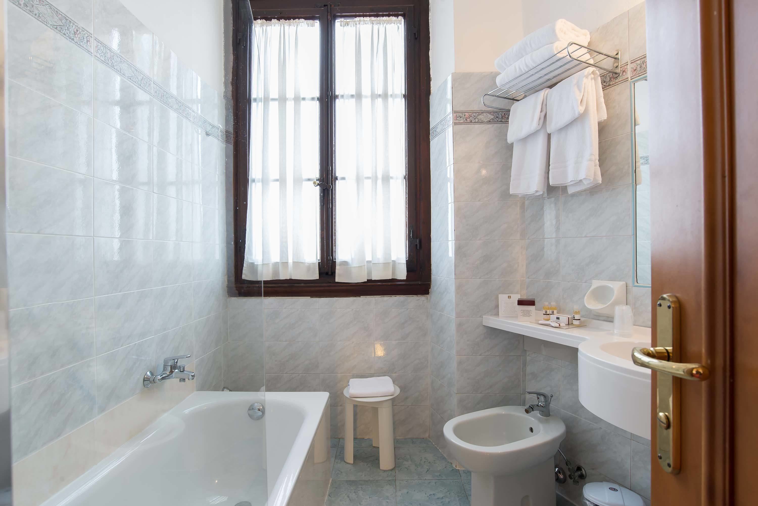 Un baño con bañera, un taburete pequeño, un bidé, un lavabo y una ventana con cortinas.