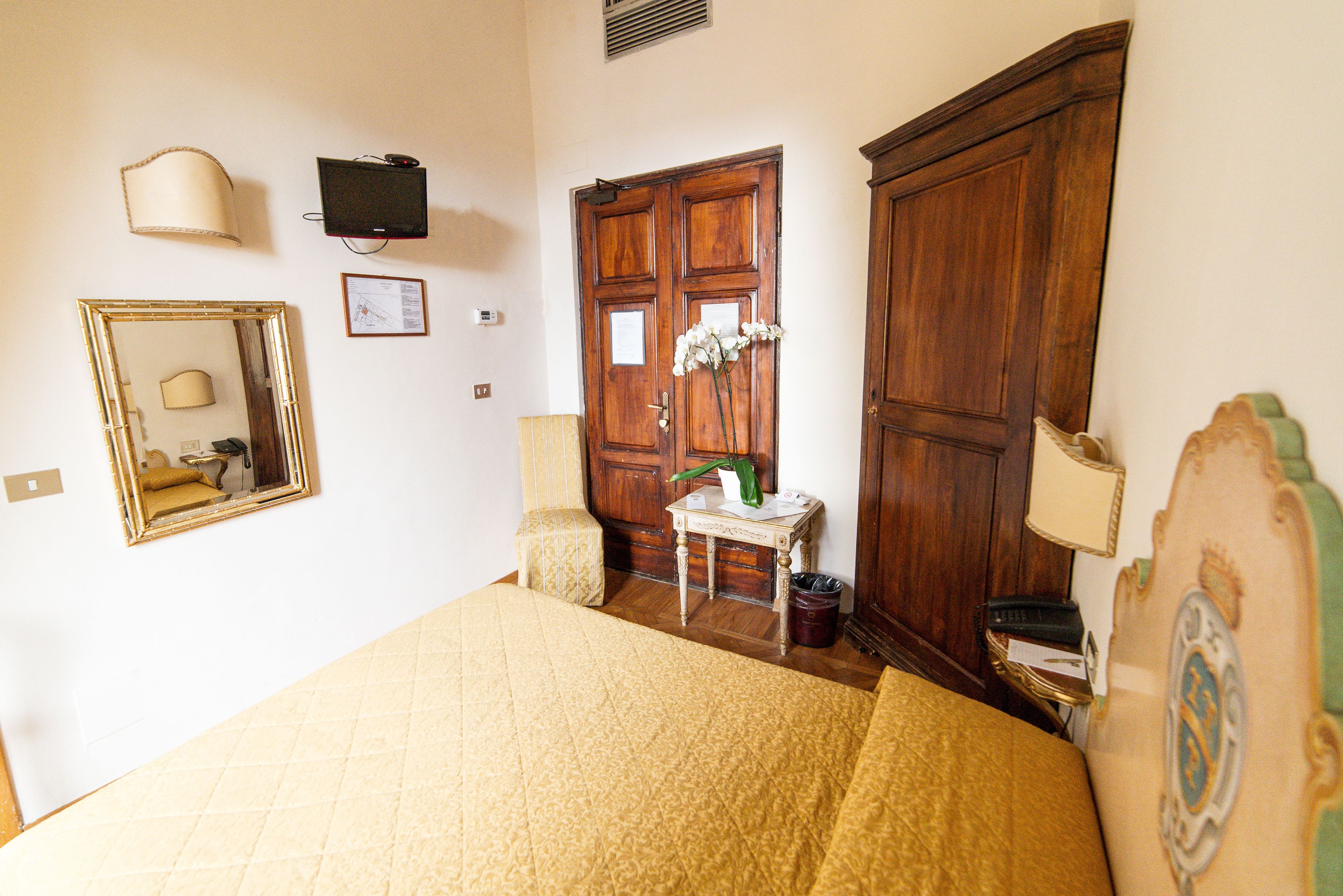 A bedroom with a large bed, a wooden wardrobe, a small bedside table with a lamp, a mirror, and a television mounted on the wall.