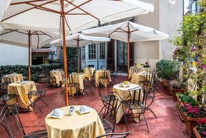 An outdoor café with round tables covered by white umbrellas, surrounded by potted plants and a red-tiled floor.
