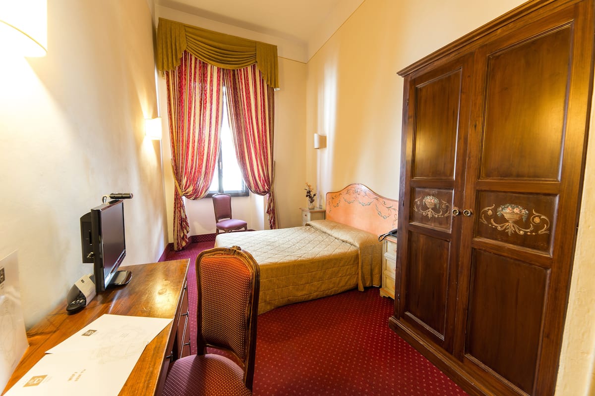 Une chambre d’hôtel comprenant un lit, un bureau avec un ordinateur, une chaise, une armoire et une fenêtre avec des rideaux.