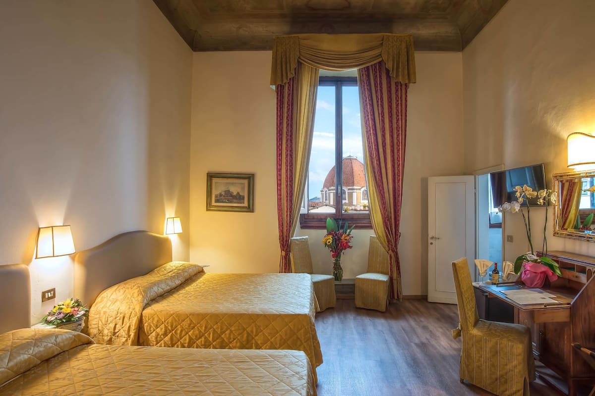 Une chambre d’hôtel avec deux lits, un bureau, une chaise, un vase de fleurs et une vue sur un dôme, visible par la fenêtre.