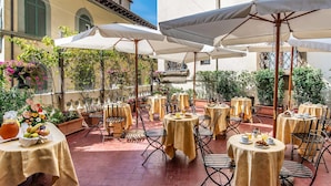 Un café en plein air avec des tables et des chaises rondes, abrité par des parasols, situé devant un bâtiment doté d’arcs en plein cintre et d’un toit recouvert de tuiles.