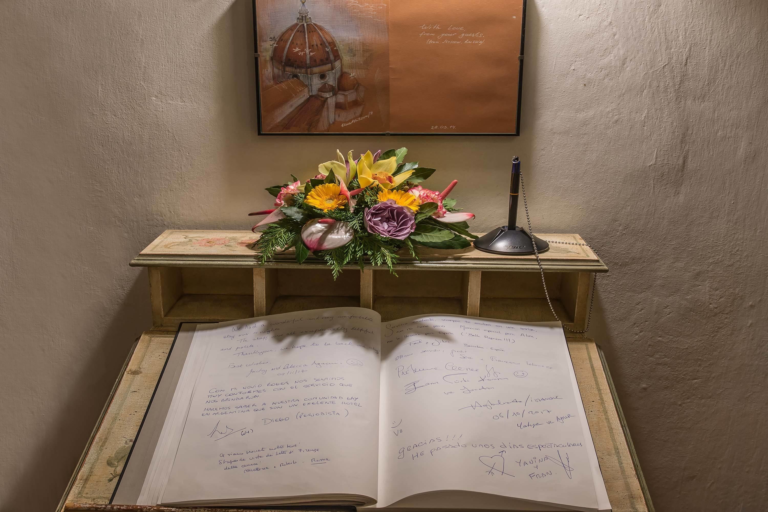 An open notebook with handwritten notes, a wooden desk with a floral arrangement, and a framed picture on the wall.