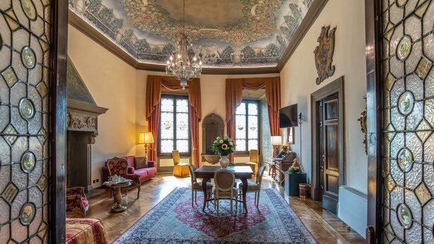 Une pièce spacieuse dotée d’un lustre, d’une cheminée, d’un coin repas avec une table et des chaises, et d’une grande fenêtre avec des rideaux.