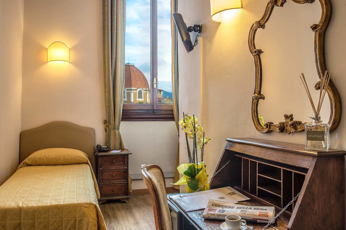 Une chambre d’hôtel avec un lit, un bureau sur lequel repose un journal, un vase avec des fleurs, un miroir et une fenêtre donnant sur un dôme.