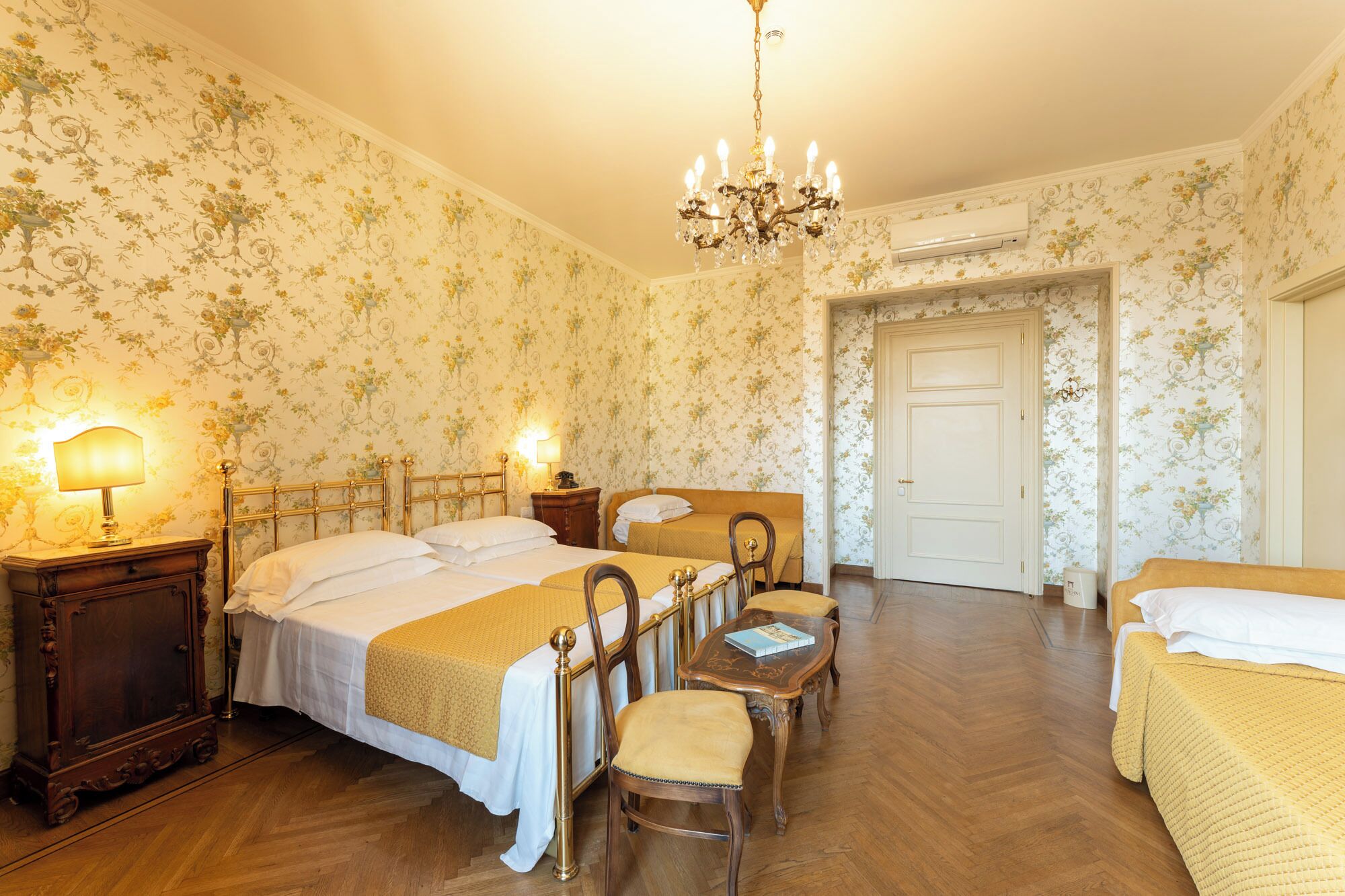 A hotel room with two beds, a wooden floor, a chandelier, and floral wallpaper.