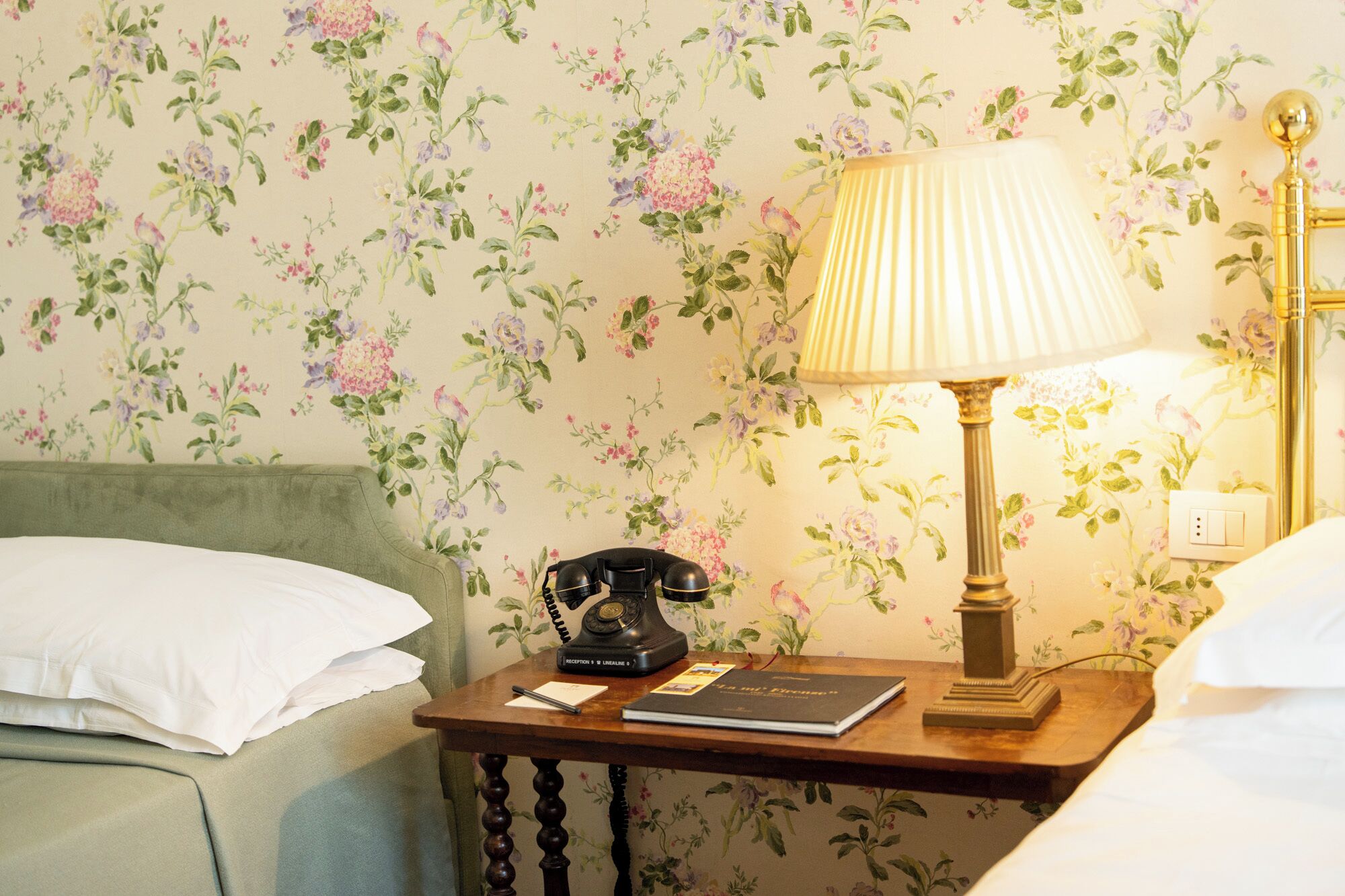 Un lit bien fait, avec une tête de lit verte, une table de chevet en bois sur laquelle se trouvent une lampe, un téléphone et un livre.