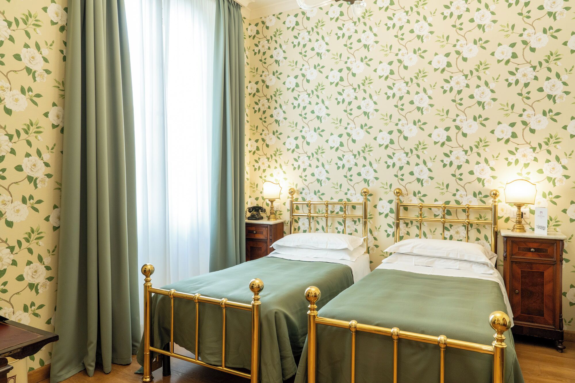 A room with two single beds, a nightstand with a lamp, a wooden dresser, and floral wallpaper.