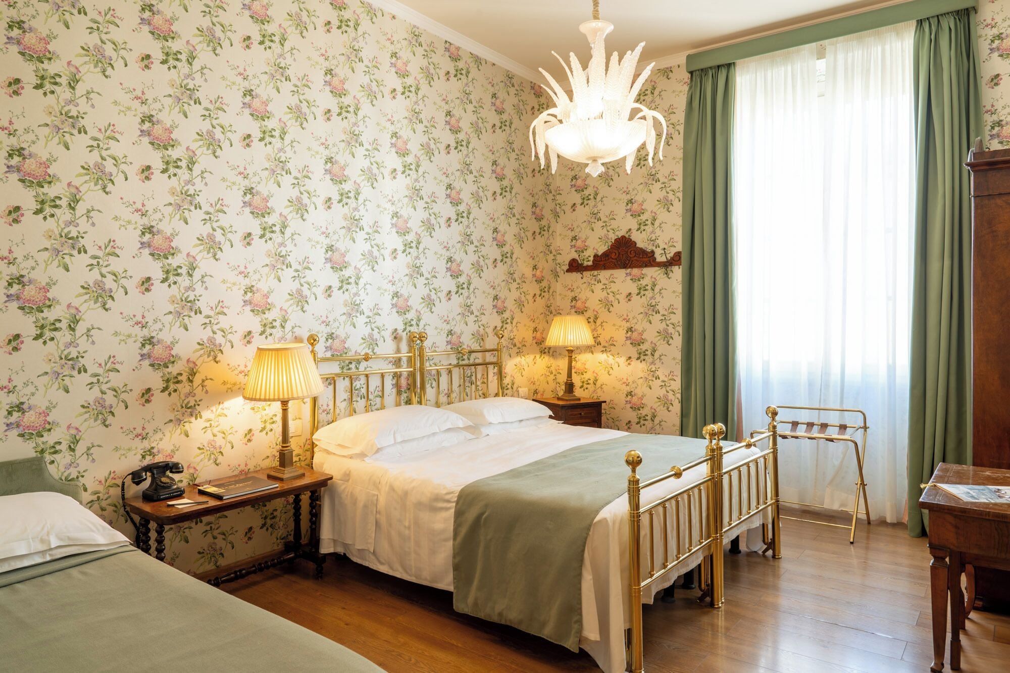 A room with floral wallpaper, a chandelier, a bed with a green bedspread, two bedside tables with lamps, and a wooden chair.