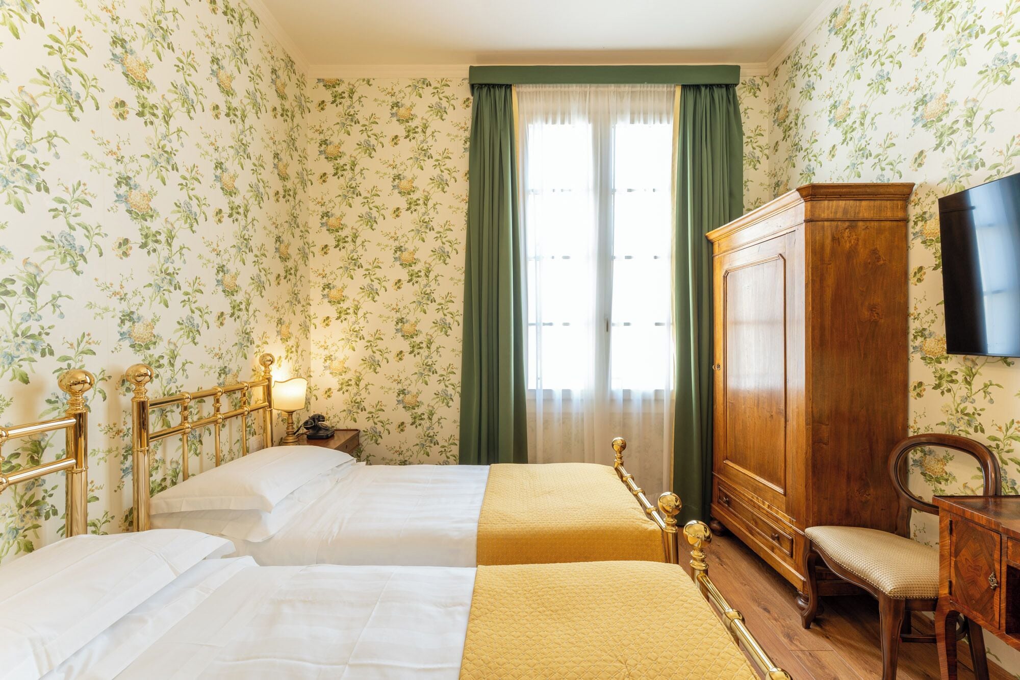 A hotel room with two beds, a wooden wardrobe, a desk with a chair, and a television on the wall.