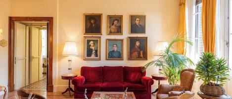Una habitación con un sofá rojo, una mesa de centro de vidrio y una pared decorada con retratos enmarcados. Hay dos lámparas sobre mesitas de noche, y se ve una planta en maceta.