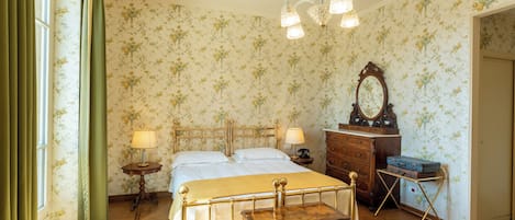 Een slaapkamer met bloemenbehang, een bed met gouden frame, twee nachtkastjes met lampen, een kaptafel, een spiegel en een koffer op een standaard.