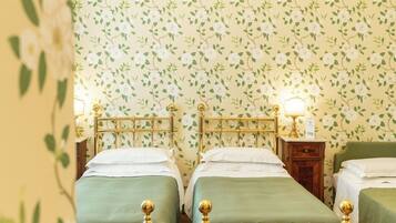 Quarto com quatro camas individuais, cada uma com colcha verde e cabeceira dourada, com um papel de parede floral ao fundo. Há duas mesas de cabeceira com luminárias, e um candelabro pendurado no teto.