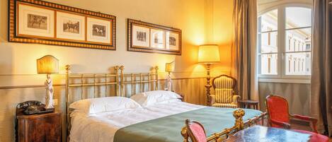 A hotel room with a bed, two bedside lamps, a chair, a table, and framed pictures on the wall.