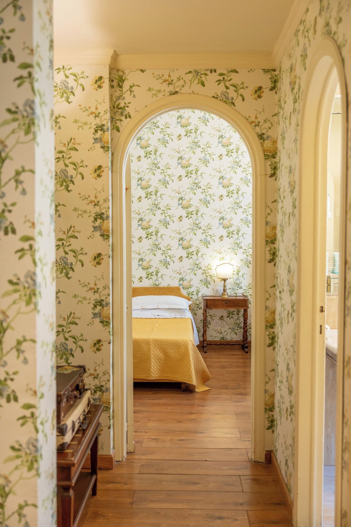 A hallway with floral wallpaper, a wooden floor, a bed with a yellow bedspread, and a nightstand with a lamp.