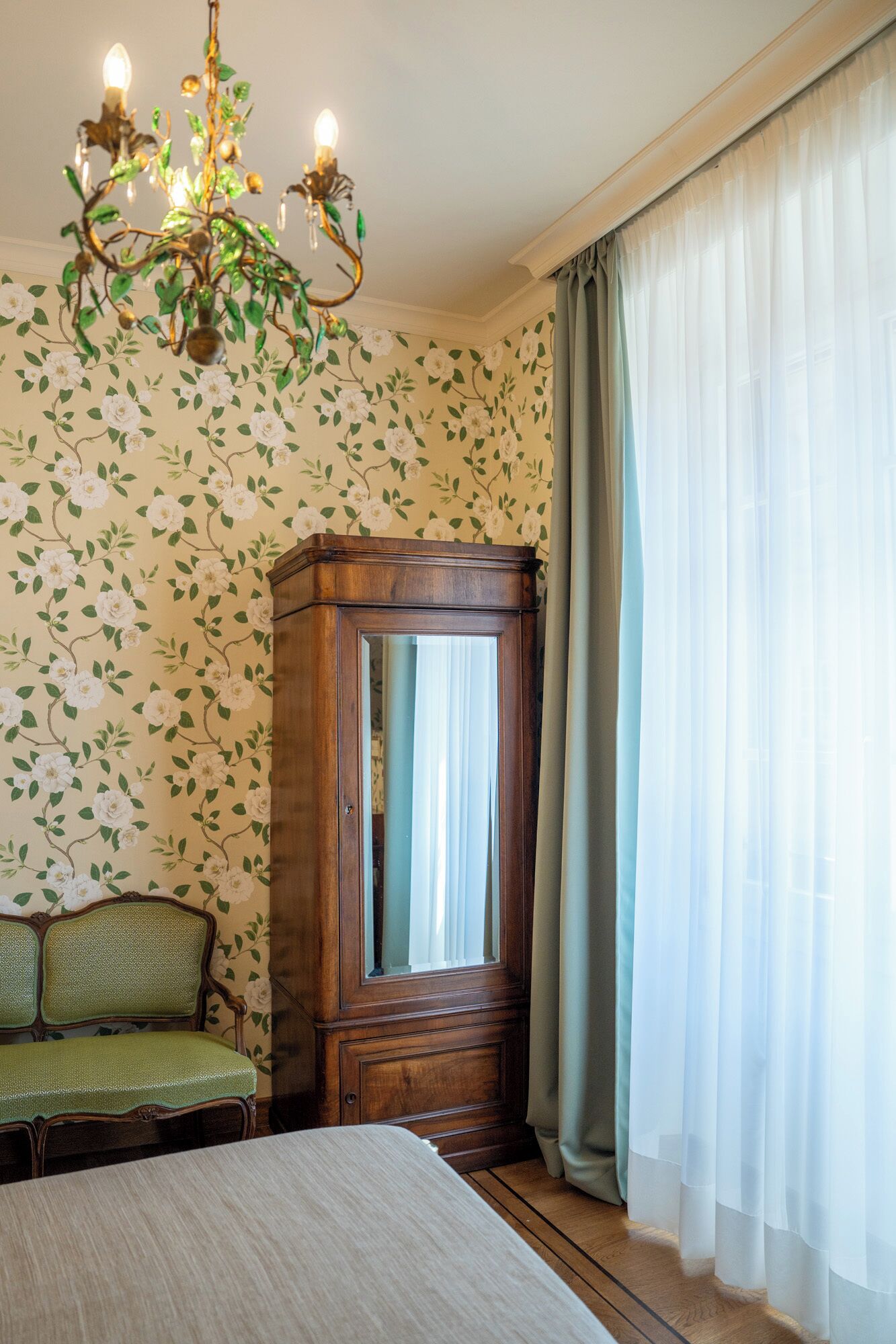 Una habitación con papel tapiz floral, un aparador de madera, un banco tapizado en verde y una cama.