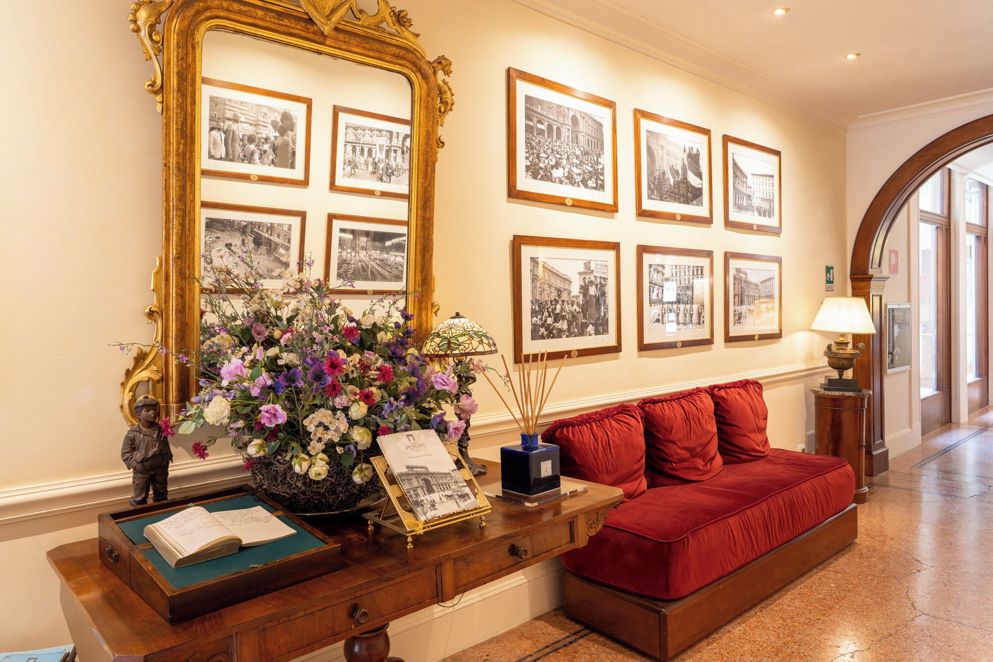 A lobby with a red sofa, a wooden console table with a vase of flowers, a large mirror, and framed black and white photographs on the wall.