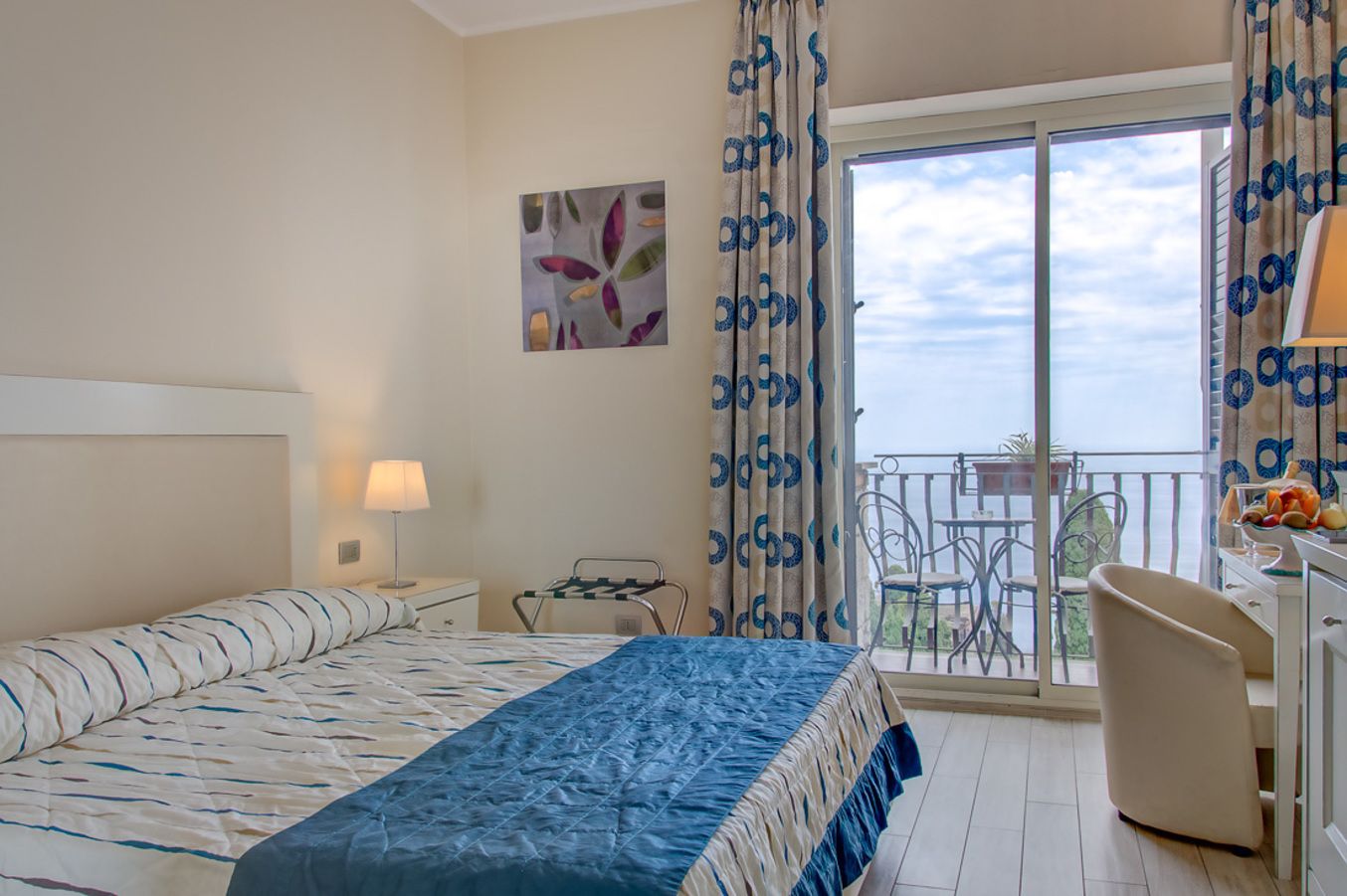 A hotel room with a bed, a desk, a chair, a balcony with a table and chairs, and a view of the sky.