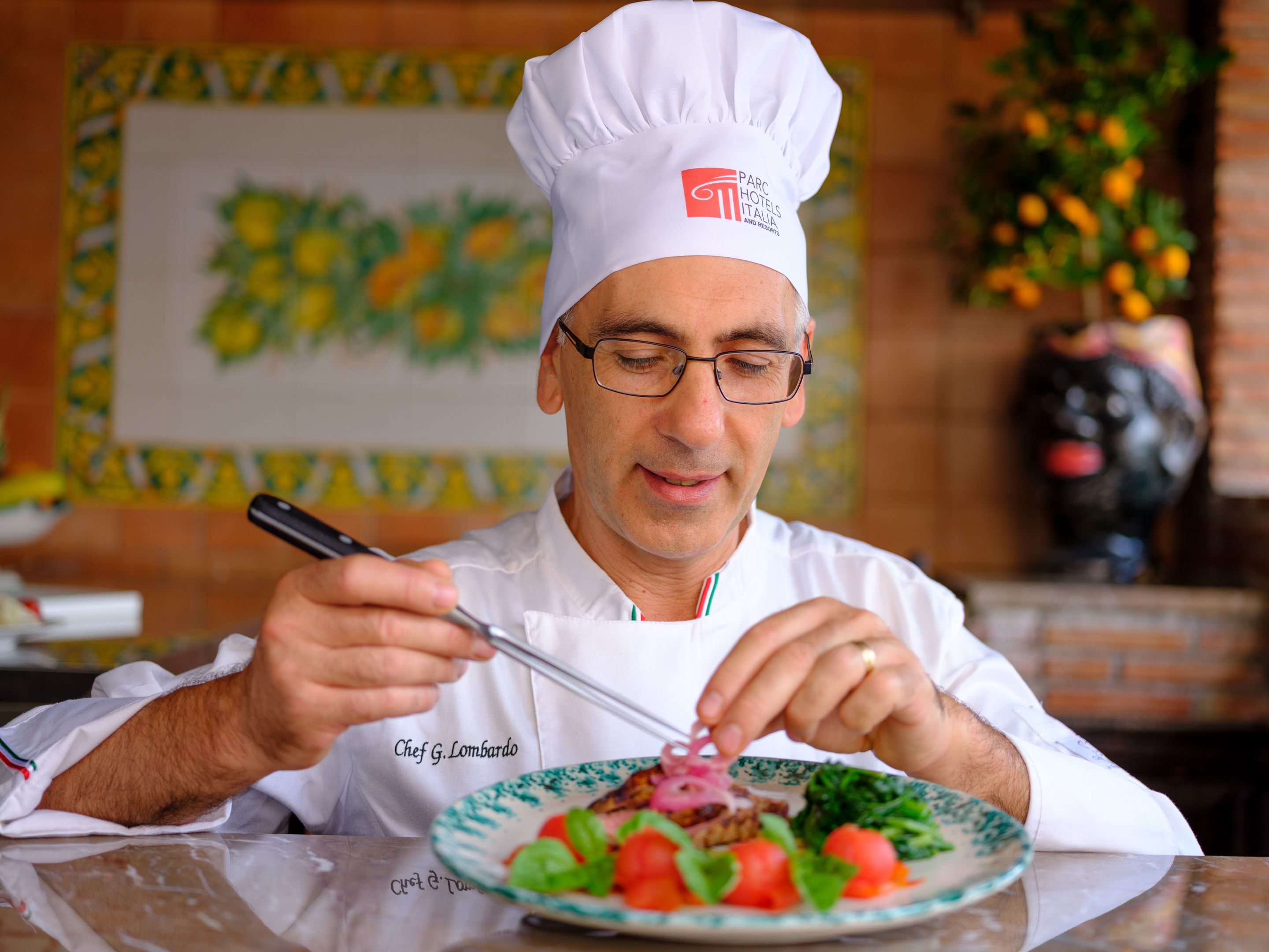A chef in a white uniform with a chef's hat, holding a fork and knife, and a plate of food in front of him.