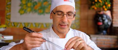 A chef in a white uniform with a chef's hat, holding a fork and knife, and a plate of food in front of him.